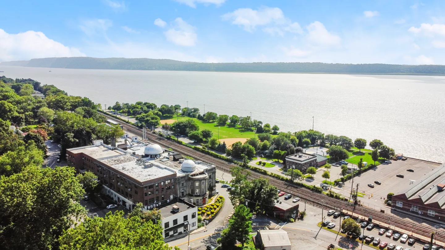 An aerial view of The Cosmopolitan building, surrounded by trees along its perimeter. The parking lot is visible, and the clear blue sky above enhances the view.