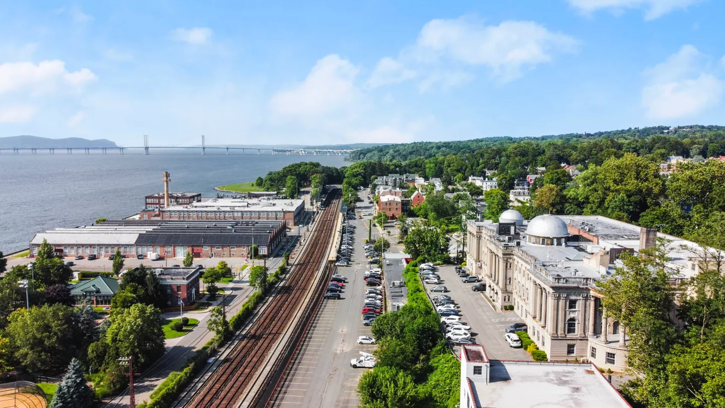 A scenic view capturing The Cosmopolitan building, the Irvington train station, and the Hudson River, all under a clear, bright blue sky.
