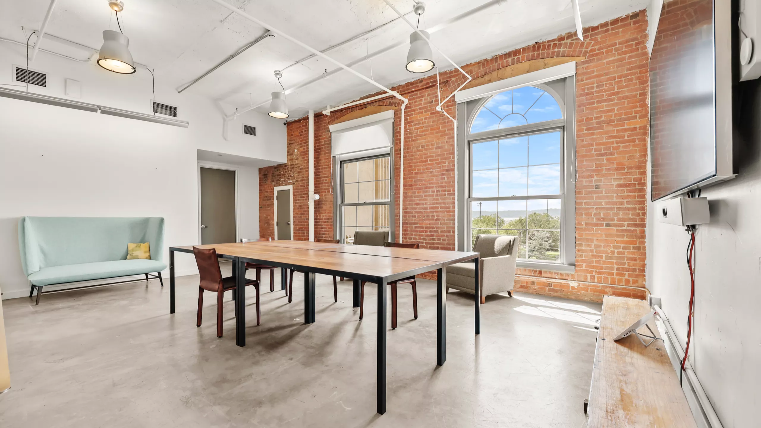 A bright, spacious office with white walls, a central meeting table, an LCD screen, and a brick wall with large windows letting in natural light. A green sofa sits in the background, complementing the modern workspace with several chairs arranged around the table.