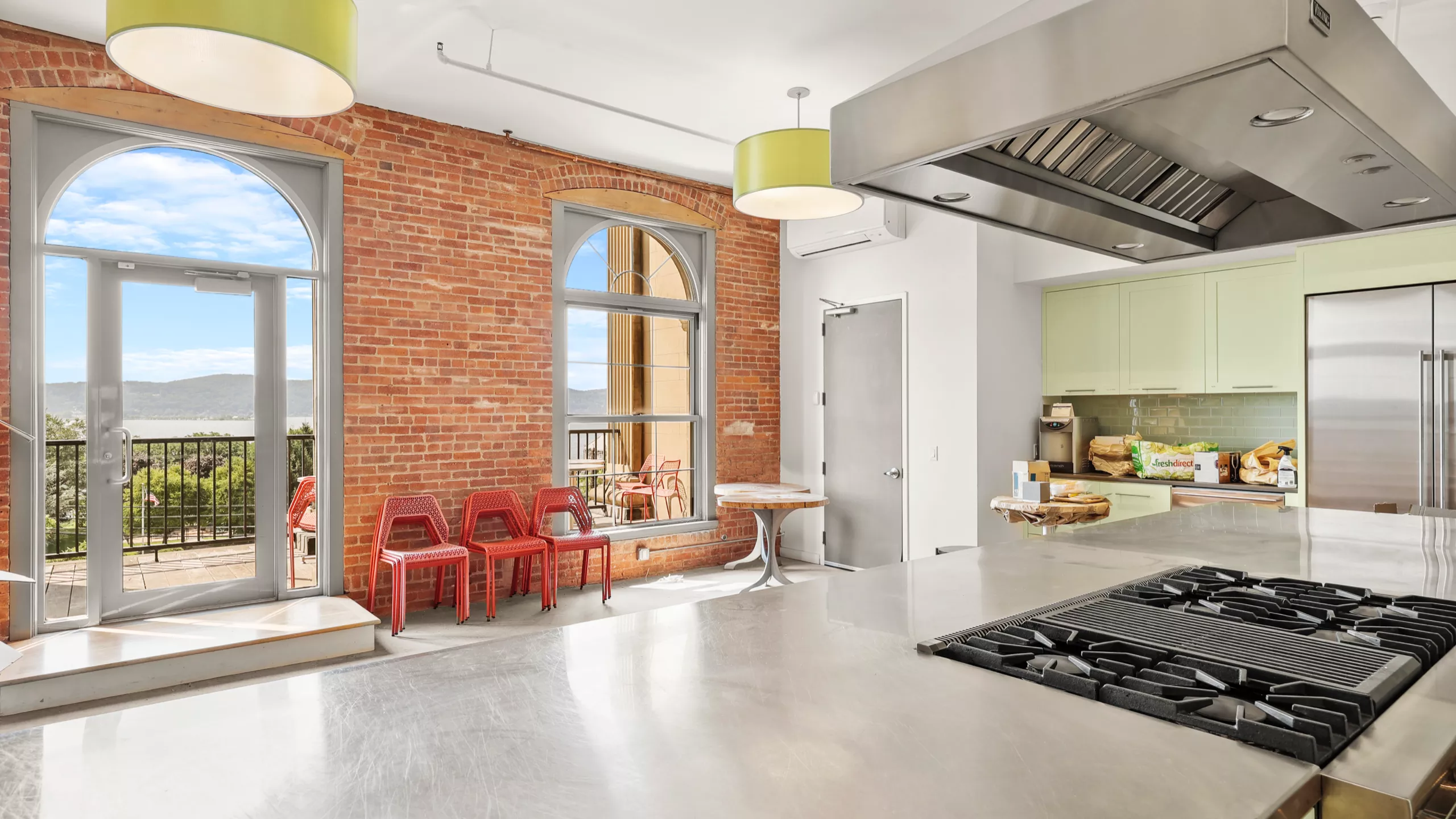 A well-equipped communal kitchen featuring cooking appliances, red chairs along the wall, and large windows offering stunning views of the Hudson River. Sunlight fills the space, creating a warm and inviting atmosphere.